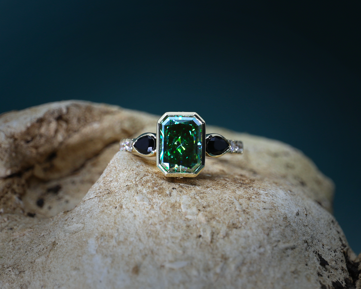 A gold ring featuring a large emerald-cut green gemstone, flanked by two small black round gemstones and tiny clear accent stones, displayed on a textured beige rock against a dark teal background.